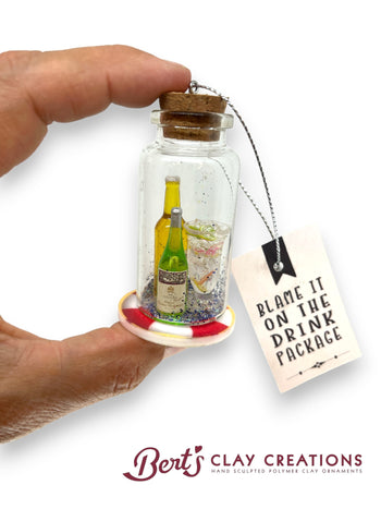 Decorative glass ornament with miniature bottles and a tag, held by a hand with 'Bert's Clay Creations' branding.